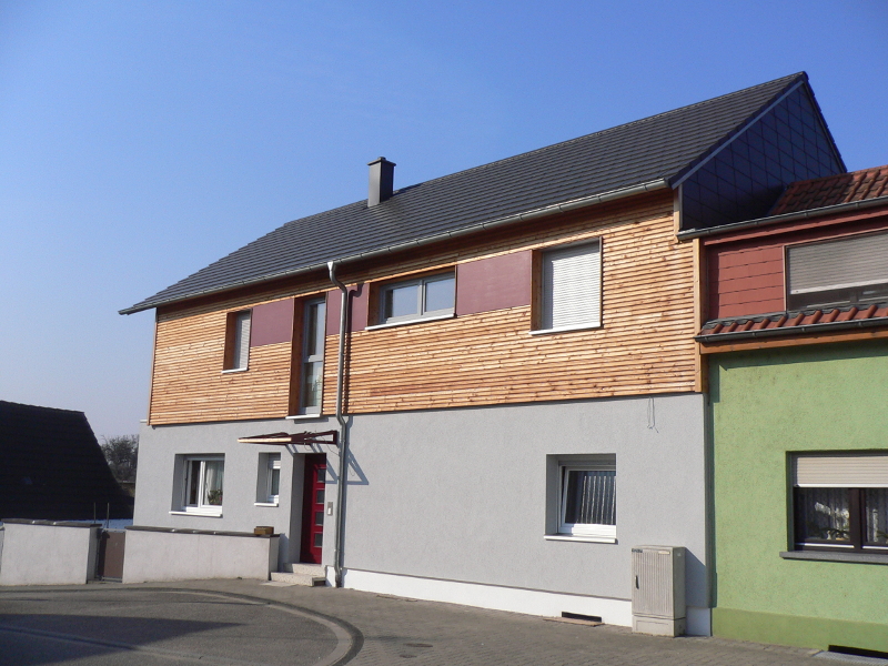Ein modernes zweistöckiges Haus mit Holzfassade, grauem Erdgeschoss und roter Tür steht neben einem grünen Gebäude unter blauem Himmel.